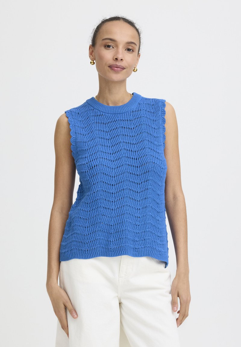 Femme portant un haut tricoté bleu sans manches et un pantalon blanc, debout devant un fond clair uni, regardant la caméra.