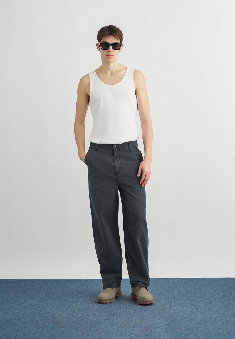 Giovane uomo con occhiali da sole neri, canottiera bianca, pantaloni larghi scuri e stivali beige in piedi con una mano in tasca davanti a uno sfondo semplice.