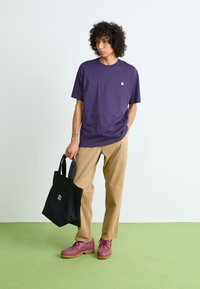 Purple cotton t-shirt, relaxed fit, short sleeves. Tan chinos, pink lace-up shoes, holding a black tote bag. Simple logo detail on shirt.