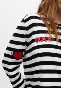 Czarnobiały sweter w paski z czerwonym napisem "LOVE" z koralików i czerwoną naszywką w kształcie serca na lewym rękawie, z motywem strzały.