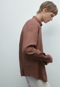 Chemise à manches longues, couleur rouille, en tissu texturé, avec un col pointu et des fentes sur les côtés pour faciliter le mouvement, portée non rentrées.