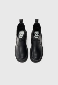 Ζευγάρι μαύρα δερμάτινα μποτάκια αστραγάλου με στρογγυλεμένες μύτες, πλευρικά φερμουάρ και χαρακτηριστικές ταινίες τράβηγματος, εμφανιζόμενα από πάνω σε λευκό φόντο.