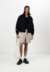 Sort bomberjakke med høj krave, beige shorts, sorte platform sko og en crèmefarvet tote bag, sat mod en hvid baggrund.