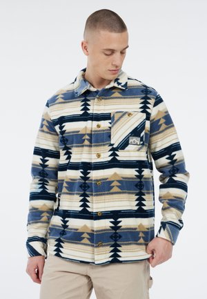 Man in een blauwe, beige en witte gebloemde fleece overhemd met een borstzak, die naar beneden kijkt terwijl hij de zoom van het shirt vasthoudt.