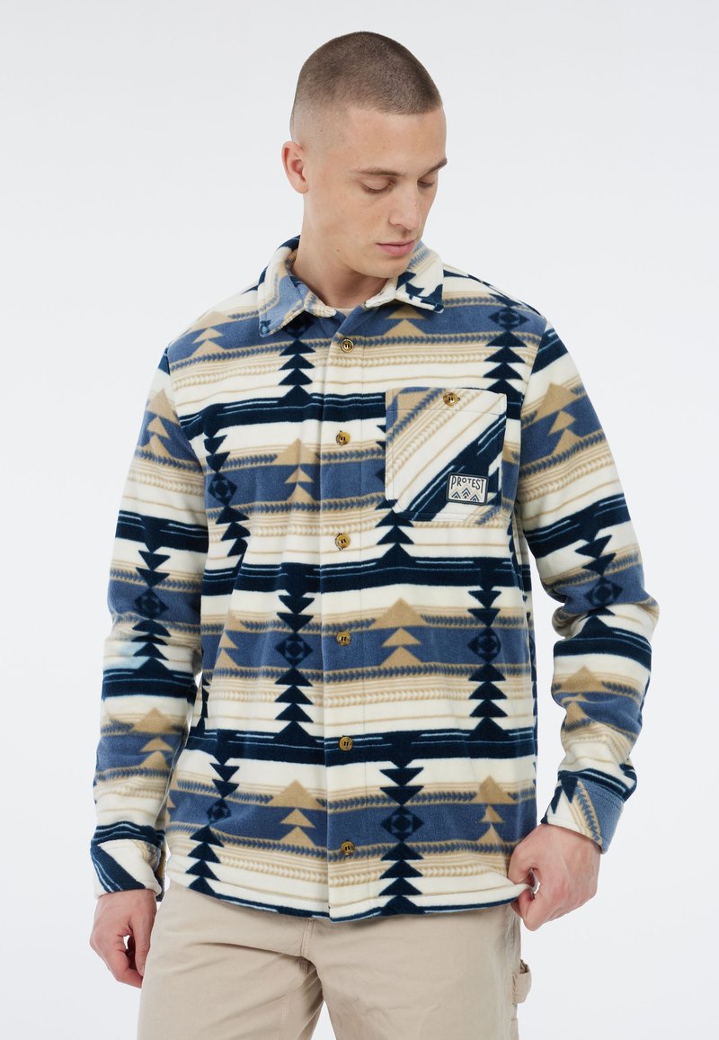 Man in een blauwe, beige en witte gebloemde fleece overhemd met een borstzak, die naar beneden kijkt terwijl hij de zoom van het shirt vasthoudt.