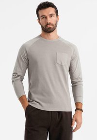 Camisa gris de manga larga con textura acanalada, que presenta un clásico escote tipo crew y un pequeño bolsillo en el pecho. Combinada con pantalones oscuros.