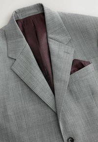Blazer gris entallado con una tela sutilmente texturizada. El interior presenta un pañuelo de bolsillo burdeos y un forro con patrón. Cierre de un solo botón.