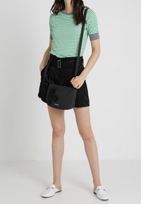 Schwarze Umhängetasche mit strukturierten Blumenakzenten, kombiniert mit einem grün-grau gestreiften T-Shirt und schwarzen Shorts. Weiße Slipper runden das Outfit ab.
