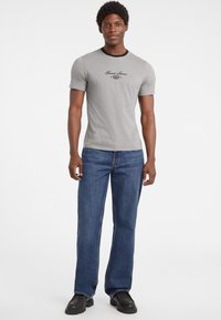 Sivý tričko s krátkym rukávom a čiernym lemom, na ktorom je logo "Guess Jeans". Skombinované s voľnými modrými džínsami a čiernymi topánkami.
