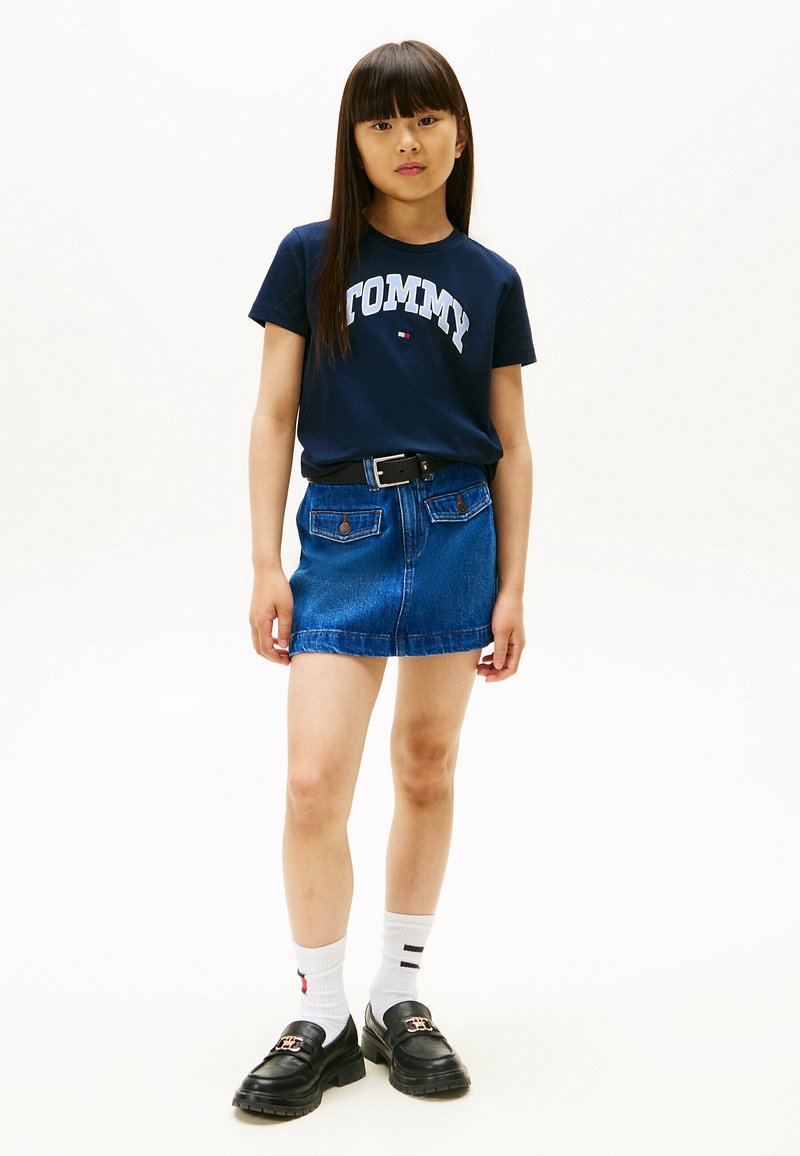 Marine t-shirt med "TOMMY" logo, denim mini nederdel med bælte, sorte loafers og hvide strømper med røde og blå accenter. Enkel design og pasform.