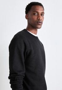Svart hettegenser med rund hals og ribbestrikkede mansjetter. Stoffet ser mykt ut, med minimal søm og en avslappet passform.