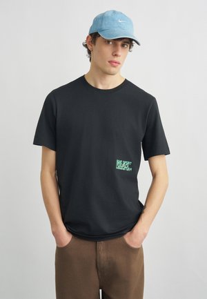 Fiatal férfi világoskék Nike sapkával, fekete Nike pólóval zöld kis logóval, és barna nadrágban, zsebeibe tett kézzel áll.