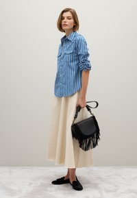 Donna in camicia a righe blu arrotolata e lunga gonna crema, che tiene una borsa frangiata nera e indossa mocassini neri, in piedi all'interno.