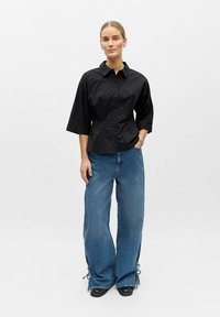 Femme debout, une main dans la poche, portant une chemise noire boutonnée à manches trois-quarts et un jean bleu à jambes larges avec des liens aux chevilles.