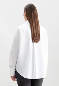 Donna con lunghi capelli castani lisci, che indossa una camicia bianca larga con bottoni e pantaloni scuri, rivolta di spalle su uno sfondo chiaro e neutro.