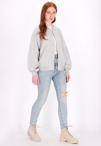 Giubbotto bomber grigio con polsini e orlo a coste, indossato su una maglietta bianca e jeans chiari strappati, abbinato a stivali beige chunky con lacci.