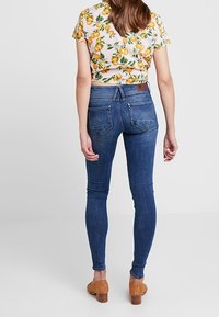 Bakre vy av en person som bär mörkblå skinny jeans med hög midja, kombinerat med en kortärmad blommig topp och bruna klacksandaler.