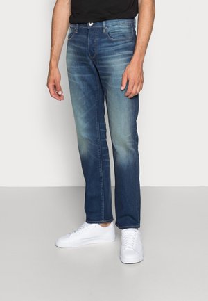 Blaue Denim-Jeans mit geradem Schnitt, mittelhoher Taille und leichter Ausbleichung. Kombiniert mit weißen Sneakern vor einem neutralen Hintergrund.