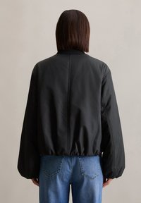 Zwarte bomberjack met een geribde kraag en elastische tailleband, met een gladde, matte textuur. Gecombineerd met lichtgewassen denim jeans.