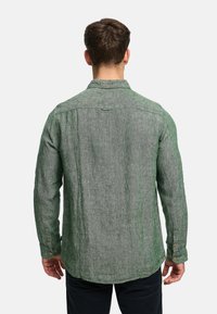 Mann mit kurzen braunen Haaren, trägt ein grünes, langärmeliges, strukturiertes Hemd und schwarze Hose, ist von hinten vor einem einfarbigen Hintergrund zu sehen.