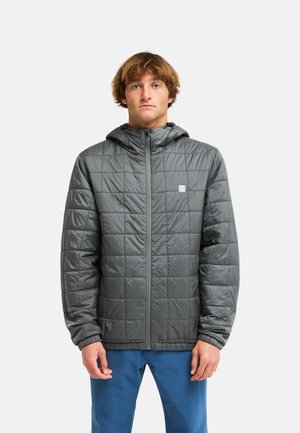 Junger Mann mit hellbraunen Haaren, der eine graue gesteppte Kapuzenjacke und blaue Hose trägt und vor einem schlichten hellen Hintergrund steht.
