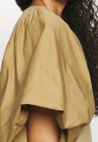 Hnědý bavlněný top s krátkými nafouklými rukávy, kulatým výstřihem a texturovaným povrchem. Tkanina má matný vzhled s jemnými záhyby.