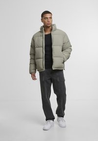 Puffjakke i olivengrønn, kviltet materiale, høy krage, glidelås. Kombinert med mørke jeans og hvite sneakers, står mot en ensfarget bakgrunn.