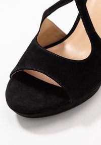 Czarne zamszowe buty na wysokim obcasie z zaokrąglonym noskiem i wyciętymi bokami, z gładkim beżowym wnętrzem i grubą platformą.