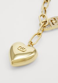 Gouden hartbedel bevestigd aan een ketting, met een gepolijst oppervlak en een reliëflogo. De schakels van de ketting zijn stevig met een sprankelende accent.