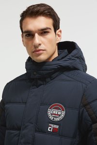 Jonge man die een donkerblauwe pufferjas met capuchon draagt, met ronde en vierkante Camp David Expedition Crew-patches op de borst.