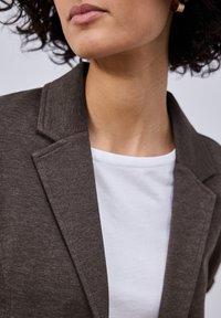 Femme portant une boucle d'oreille en or en forme d'anneau, un blazer marron sur une chemise blanche, avec des cheveux bouclés et une couleur de lèvres neutre, montrée de la mâchoire aux épaules.
