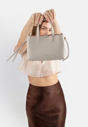 Donna che tiene una borsa in pelle beige con doppi manici davanti al viso, indossando una blusa corta e una gonna di raso marrone.