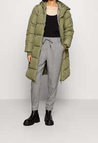 Olivgrön puffjacka med huva, svart linne, grå hundtandsmönstrade byxor, och svarta ankelskoot med tjock sula.