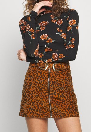 Mujer con blusa negra de manga larga con estampado floral, combinada con una falda de pana marrón con estampado de leopardo, que incluye una cremallera frontal y un cinturón con hebilla plateada.
