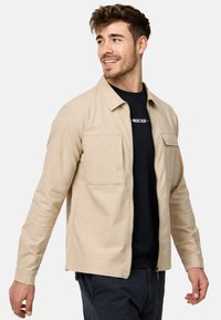 Veste zippée beige avec deux poches poitrine, design à col et manches longues. Portée par-dessus une chemise noire, présentant une texture de tissu lisse.