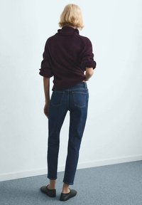 Osoba s krátkými blond vlasy, oblečená v tmavě fialovém svetru, modrých džínách a černých botách, stojící zády k přední straně.