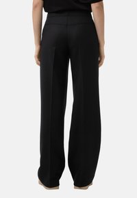 Osoba oblečená ve volných černých kalhotách a černém topu, stojící zády k fotoaparátu na čistě bílém pozadí.