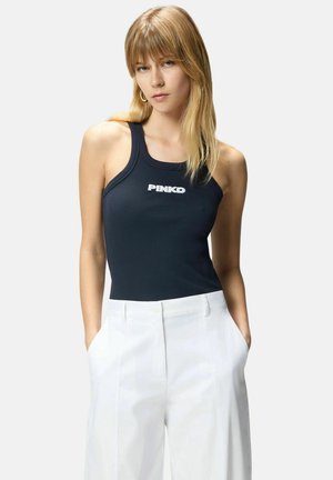 Donna bionda con top nero senza maniche con scritta "PINKO" e pantaloni bianchi a vita alta, mani in tasca, sfondo neutro.