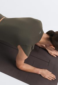 Person i olivengrøn skjorte med "Oysho"-logo, der ligger på maven på en mørk yogamåtte med armene strakt fremad.