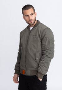 Hombre con cabello corto y barba, vistiendo una chaqueta bomber verde con bolsillo con cremallera en el brazo, mirando hacia la izquierda sobre un fondo blanco.