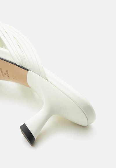 Άσπρο δερμάτινο mule με πλισέ επάνω μέρος και μαύρη μύτη στη καμπυλωτή μεσαίου ύψους φτέρνα, εμφανίζεται από πλαϊνή γωνία.