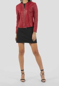 Veste en cuir rouge à col rond, fermeture zippée et coutures détaillées, portée sur une mini-robe noire et associée à des sandales noires à lanières.