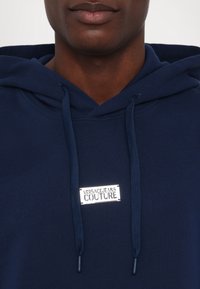 Marineblauer Hoodie aus weichem Stoff mit einer Kängurutasche vorne, versehen mit einem silbernen Logetikett und Kordelzügen. Einfaches und modernes Design.