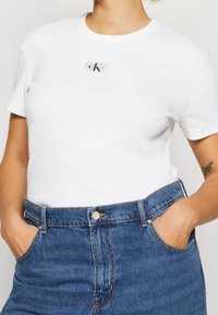 T-shirt bianca in cotone con maniche corte e collo rotondo, caratterizzata da un piccolo etichetta nera con il logo "CK" sul petto.