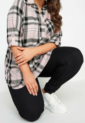 Persona que lleva una camisa de cuadros rosa y gris, pantalones negros y zapatillas blancas, arrodillada con las manos apoyadas en una rodilla contra un fondo liso.
