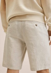 Beige pellavashortsit suoran leikkauksen kanssa, joissa on kaksi takataskua napillisia sulkemisia varten ja sileä pinta, näkyvissä kyynärpään keskikohdalla.