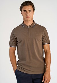 Brun pikétröja med kort ärm, med en zip-kant på kragen och marinblå och vita randiga detaljer på kragen och ärmarna.