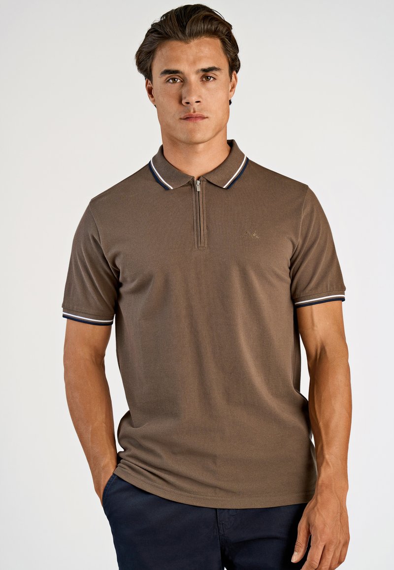 Brun pikétröja med kort ärm, med en zip-kant på kragen och marinblå och vita randiga detaljer på kragen och ärmarna.