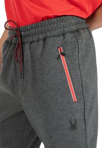 Graue Sporthose aus strukturierter Stoff, elastischem Bund, ropem Kordelzug, seitlichem Reißverschluss mit rot-weißen Akzenten und Spinnenlogo.