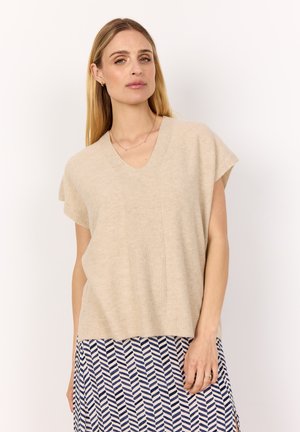 Beige stickad tröja med V-ringning, korta ärmar och ribbade detaljer. Modellen bär den över en mönstrad kjol med blå och vita zigzag-mönster.